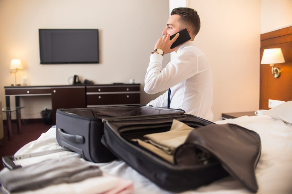 Θεματική εικόνα για: τι πρέπει να έχει ένα δωμάτιο ξενοδοχείου. Επισκέπτης σε κρεβάτι μιλά στο κινητό, με ανοιχτή βαλίτσα δίπλα.